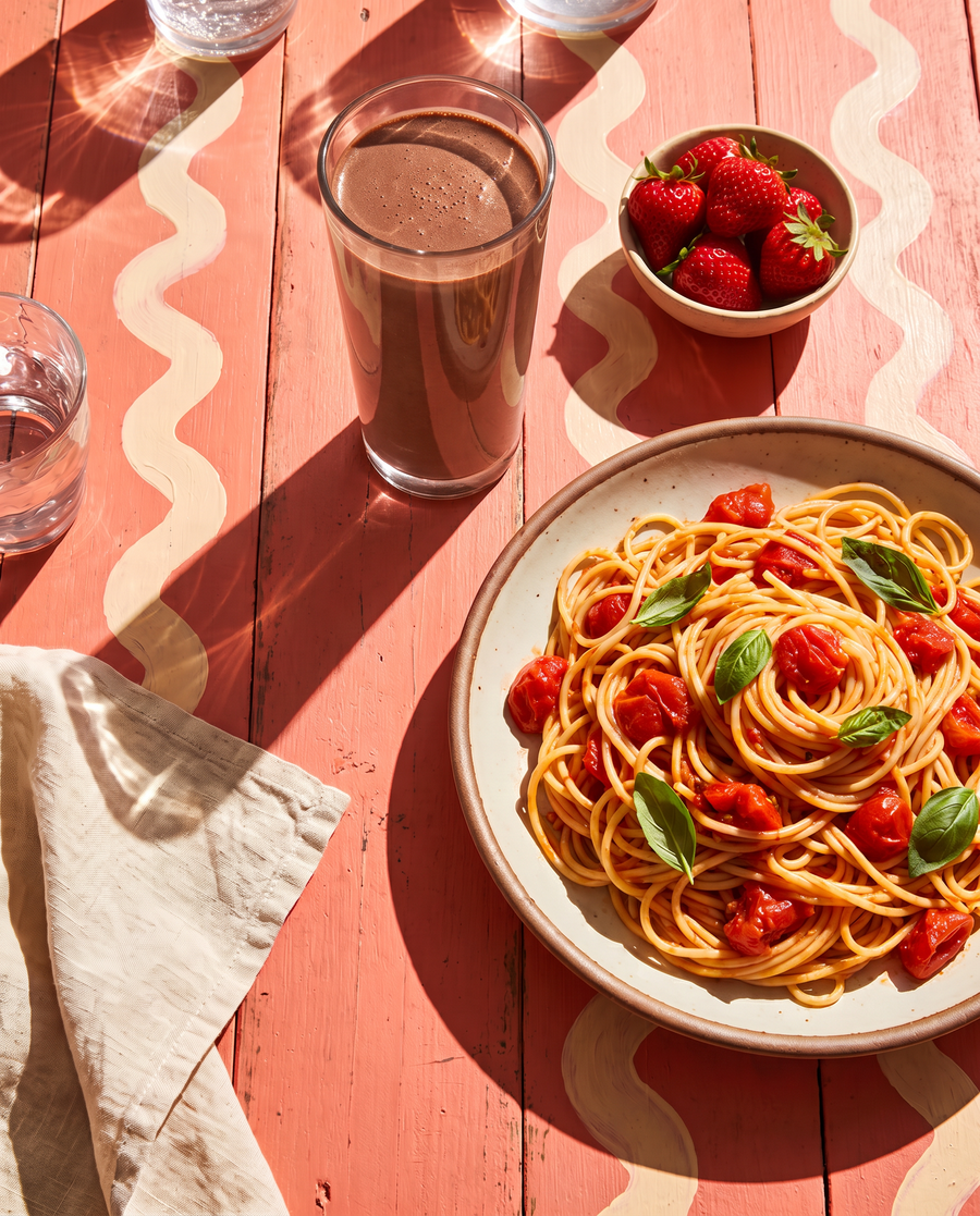 A sunny table with a plate of tomato pasta, a chocolate shake and strawberries
