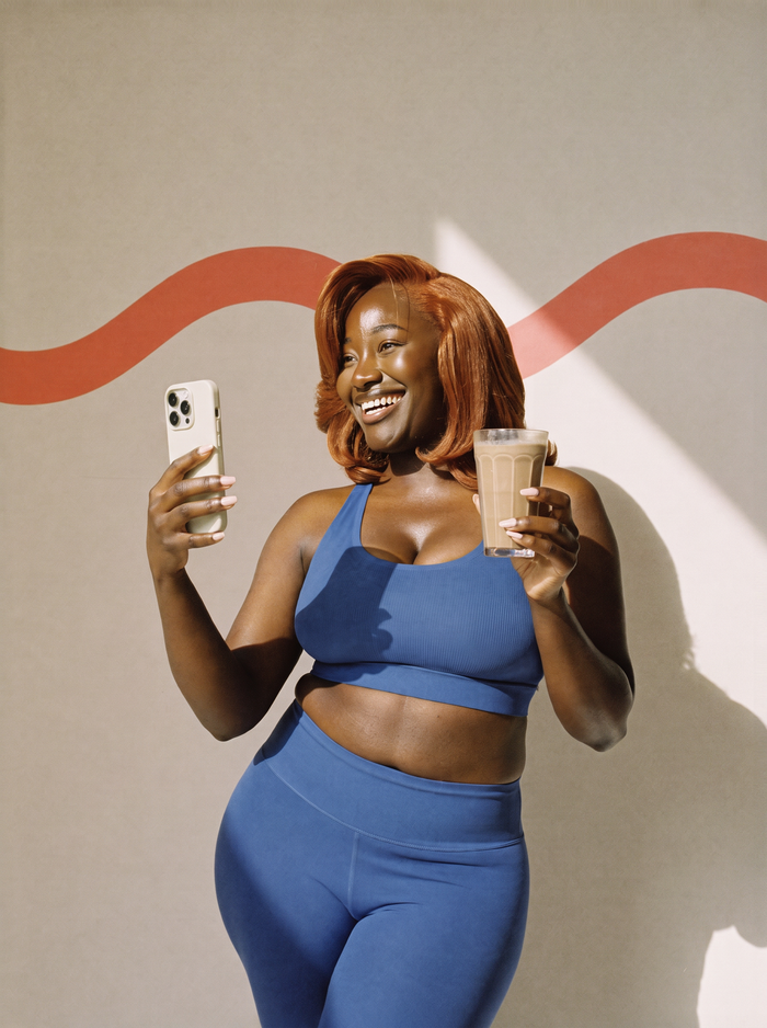 AI illustration: a woman in blue sportswear laughing as she films herself, a glass of chocolate shake in her hand