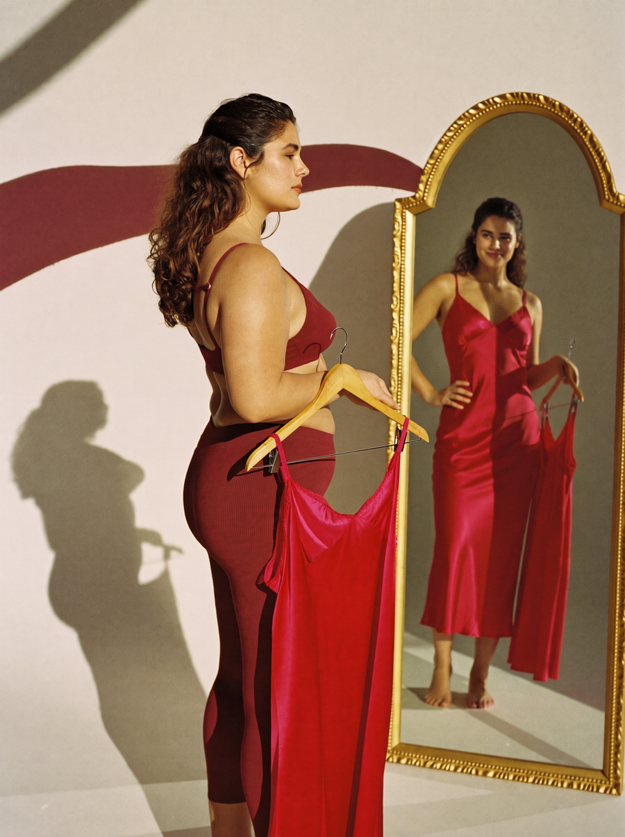 AI illustration: a woman in bordeaux sportswear holds a red satin dress, and her slimmer reflection wears it with confidence